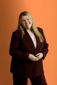 A woman with long blonde hair, wearing a maroon suit and light pink top, smiles while standing against an orange background.