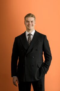 A man in a dark suit, white shirt, and polka-dot tie stands against an orange background, smiling with one hand in his pocket.