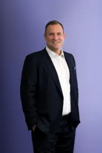 A man in a navy suit and white shirt stands smiling against a purple background, with his left hand in his pocket.