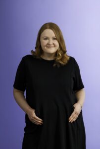 A woman with wavy, shoulder-length red hair stands against a light purple background, smiling gently. She is wearing a black dress with short sleeves and has her hands in her pockets.