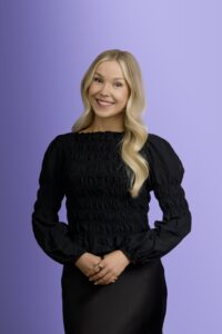 A smiling woman with long blonde hair wearing a black, long-sleeved blouse stands against a light purple background, with her hands gently clasped in front of her.