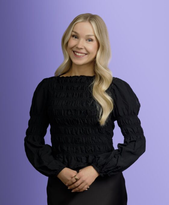 A smiling woman with long blonde hair wearing a black, long-sleeved blouse stands against a light purple background, with her hands gently clasped in front of her.