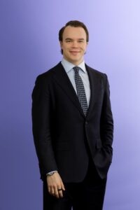 A man in a dark suit, light blue shirt, and patterned tie stands against a plain light purple background, smiling slightly with one hand in his pocket.