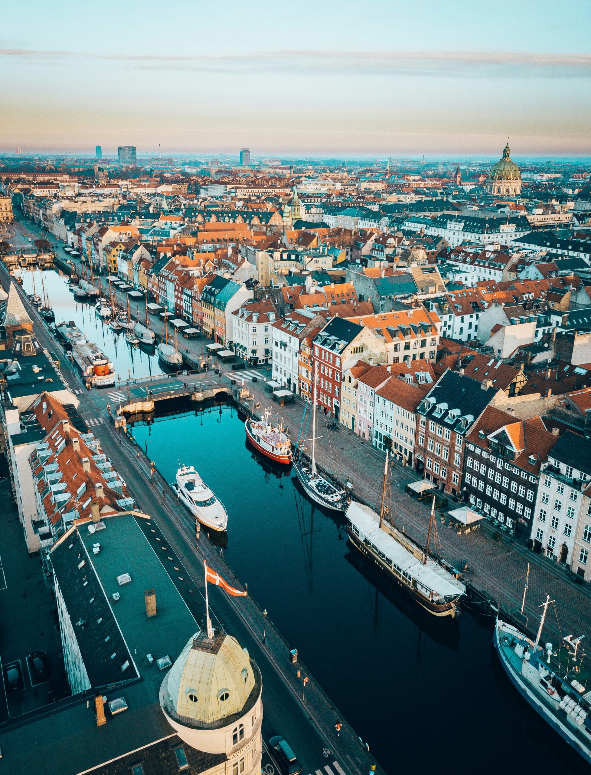Aerial view of colorful buildings and boats along a canal in the Nyhavn district of Copenhagen, Denmark, with the cityscape and dome of Frederik’s Church visible in the background under a clear sky.