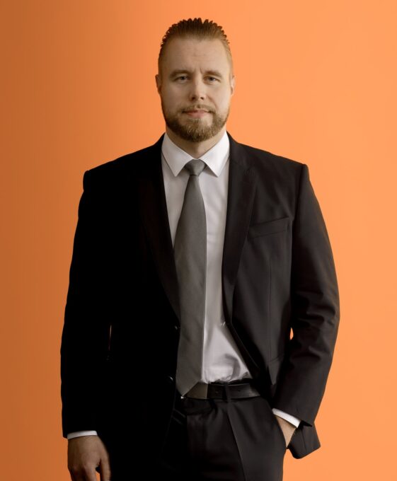 A man with light brown hair and a beard, wearing a black suit, white shirt, and gray tie, stands against an orange background with one hand in his pocket.