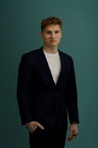 A young man with short, light brown hair wearing a dark suit jacket over a white t-shirt stands against a plain teal background, looking at the camera with a neutral expression and one hand in his pocket.