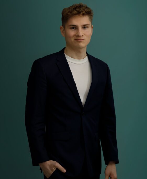 A young man with short, light brown hair wearing a dark suit jacket over a white t-shirt stands against a plain teal background, looking at the camera with a neutral expression and one hand in his pocket.