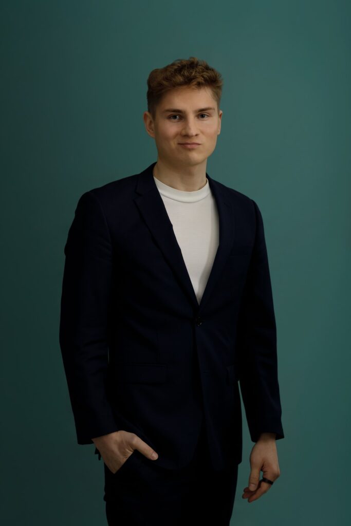 A young man with short, light brown hair wearing a dark suit jacket over a white t-shirt stands against a plain teal background, looking at the camera with a neutral expression and one hand in his pocket.