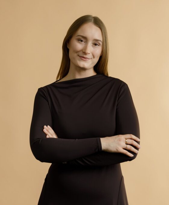A woman with straight, light brown hair wearing a long-sleeved black dress stands against a beige background, smiling softly with her arms crossed.