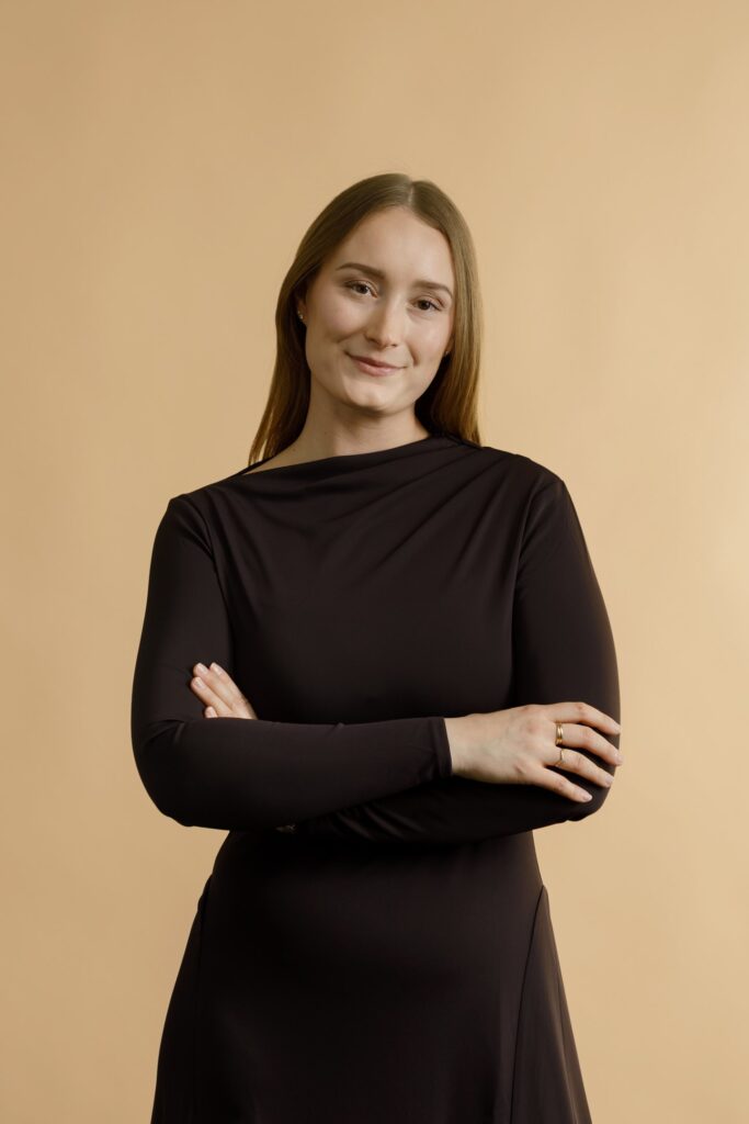 A woman with straight, light brown hair wearing a long-sleeved black dress stands against a beige background, smiling softly with her arms crossed.