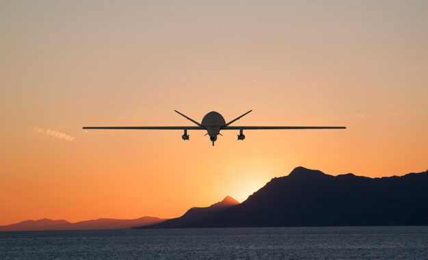 A drone flies over the ocean at sunset, silhouetted against an orange sky with mountains in the background.