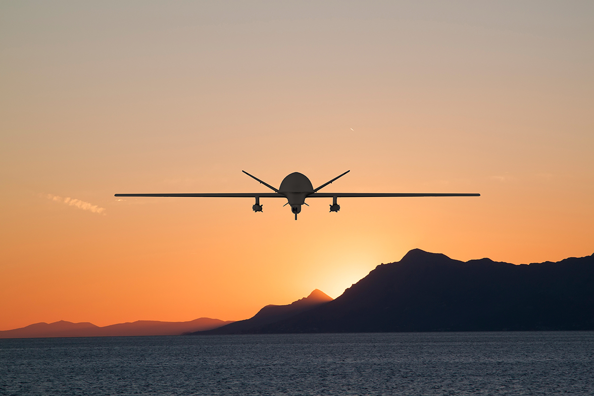 A drone flies over the ocean at sunset, silhouetted against an orange sky with mountains in the background.
