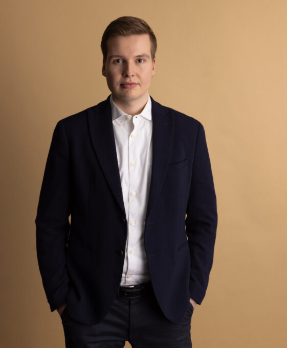 A young man stands against a plain beige background, wearing a dark blazer over a white shirt. He has short light brown hair and is looking directly at the camera with a neutral expression, hands in his pockets.