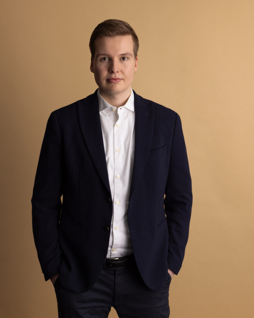 A young man stands against a plain beige background, wearing a dark blazer over a white shirt. He has short light brown hair and is looking directly at the camera with a neutral expression, hands in his pockets.