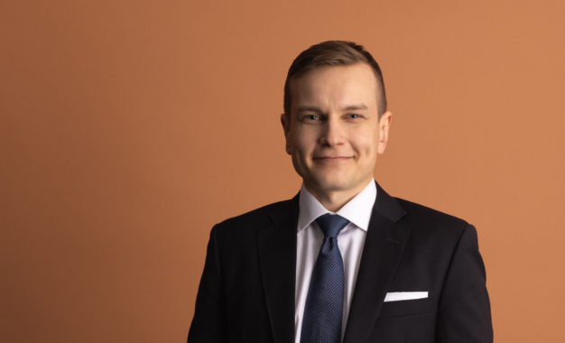A man in a black suit, white shirt, and blue patterned tie smiles at the camera against a rust-colored background.
