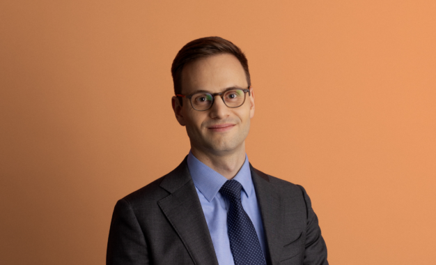 A man wearing glasses, a gray suit, blue shirt, and navy tie smiles slightly while standing against a plain peach-colored background.
