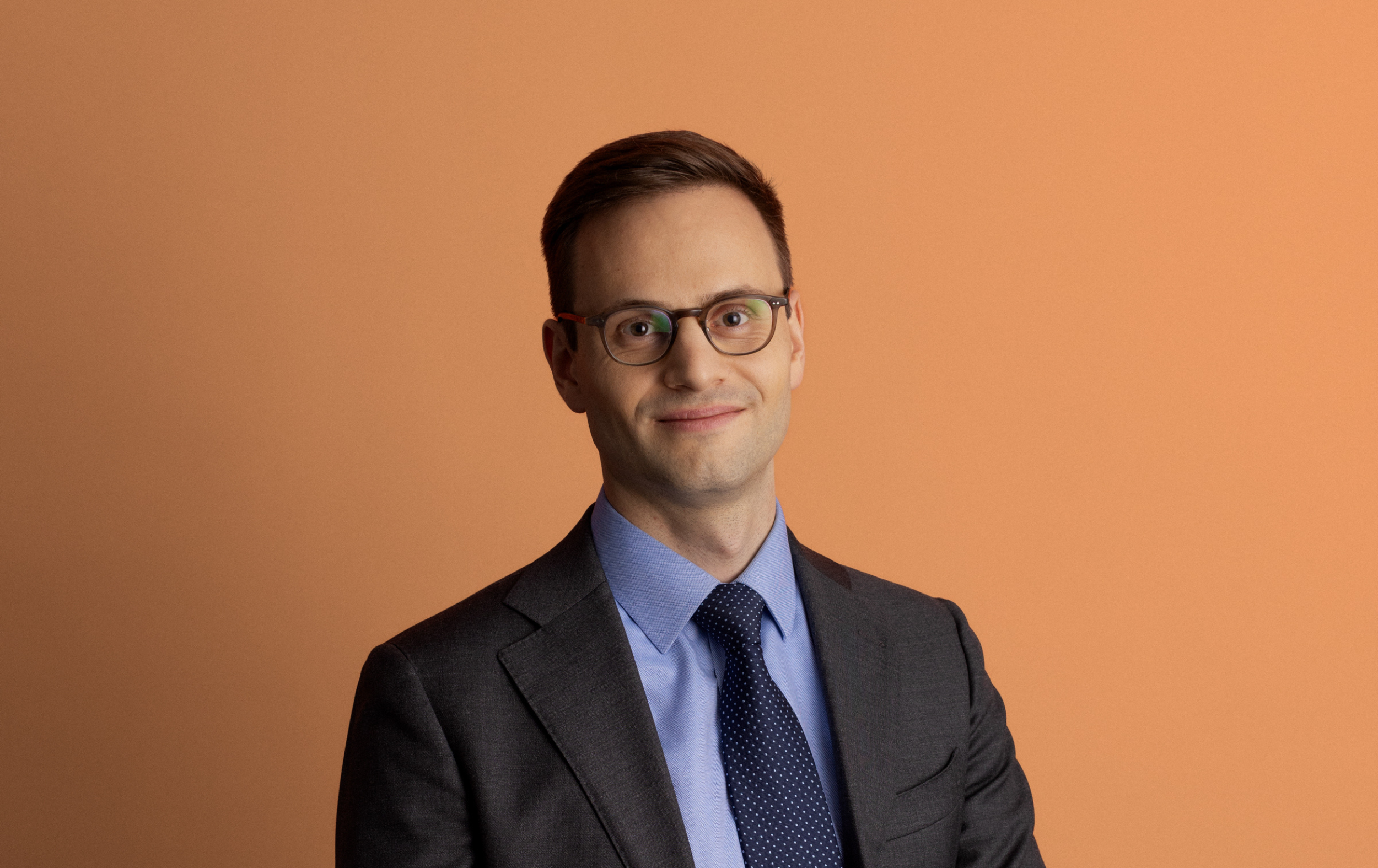 A man wearing glasses, a gray suit, blue shirt, and navy tie smiles slightly while standing against a plain peach-colored background.