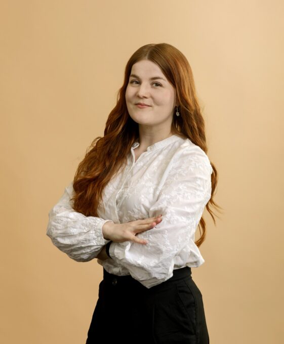 A person with long red hair stands against a beige background, wearing a white blouse and black pants, smiling gently with arms crossed.
