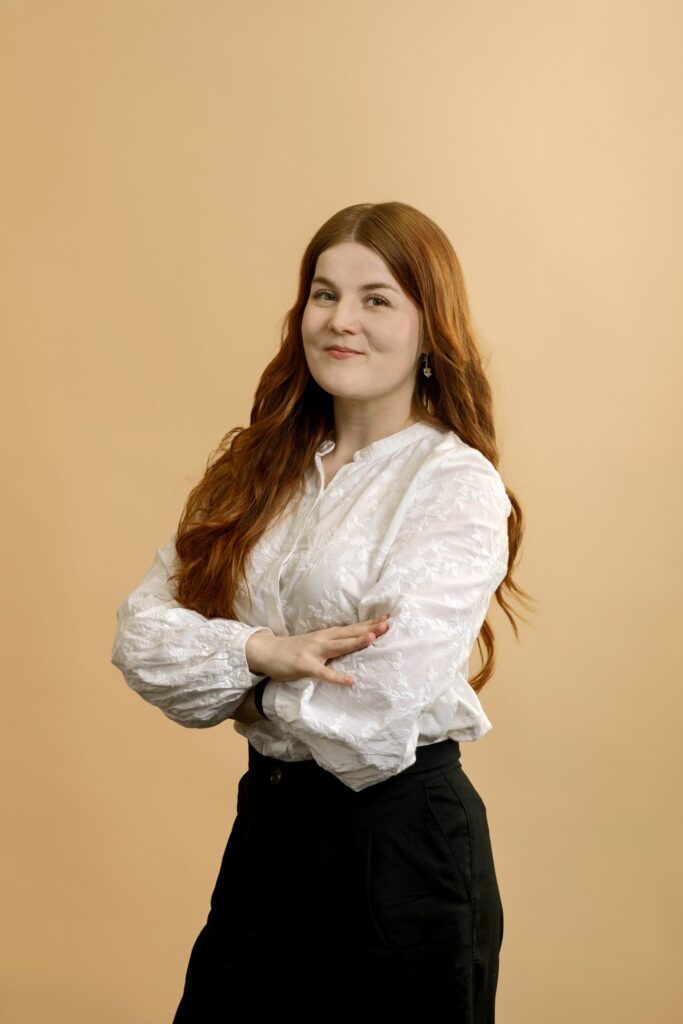 A person with long red hair stands against a beige background, wearing a white blouse and black pants, smiling gently with arms crossed.