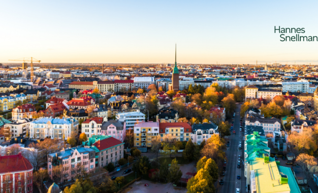 Aerial view of a colorful cityscape with various buildings, trees, and a church spire at sunrise or sunset. The skyline stretches into the distance. The words Hannes Snellman appear in the sky area on the right.