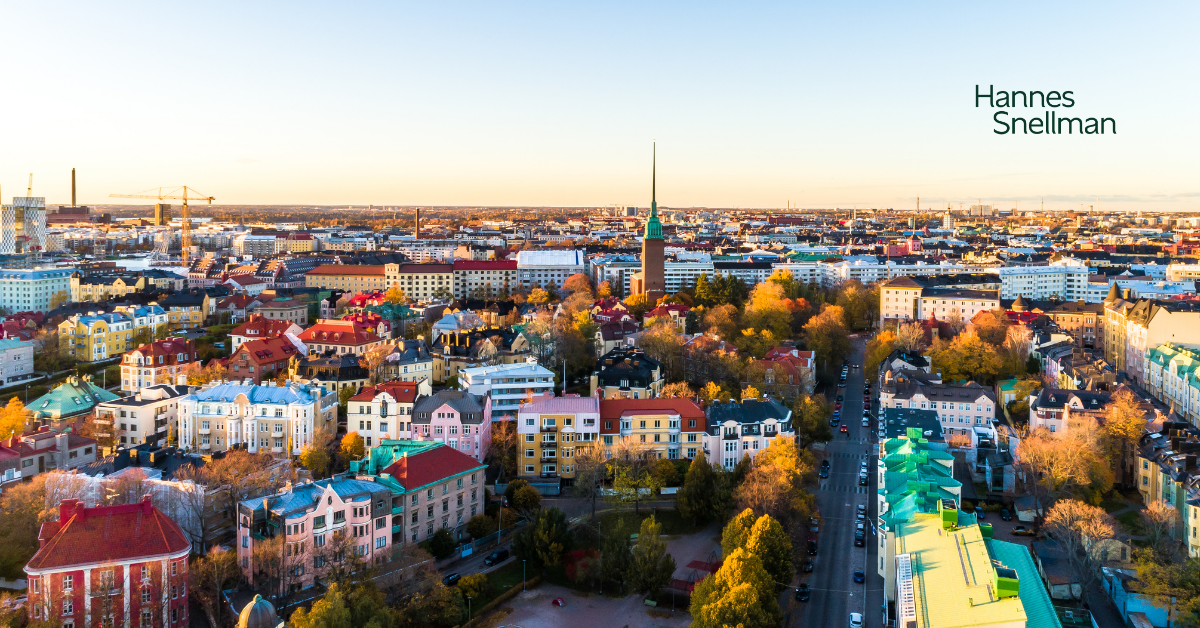 Aerial view of a colorful cityscape with various buildings, trees, and a church spire at sunrise or sunset. The skyline stretches into the distance. The words Hannes Snellman appear in the sky area on the right.