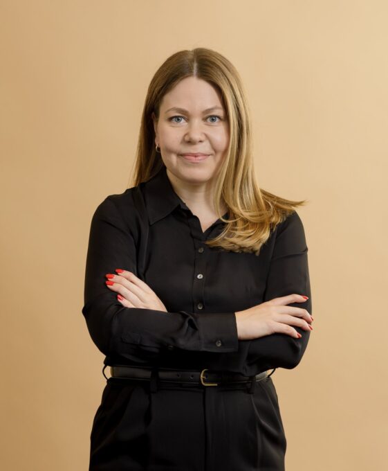 A woman with long blonde hair wearing a black button-up shirt and black pants stands with her arms crossed, smiling slightly, against a plain beige background.
