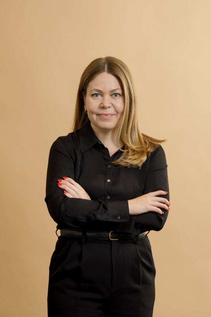 A woman with long blonde hair wearing a black button-up shirt and black pants stands with her arms crossed, smiling slightly, against a plain beige background.
