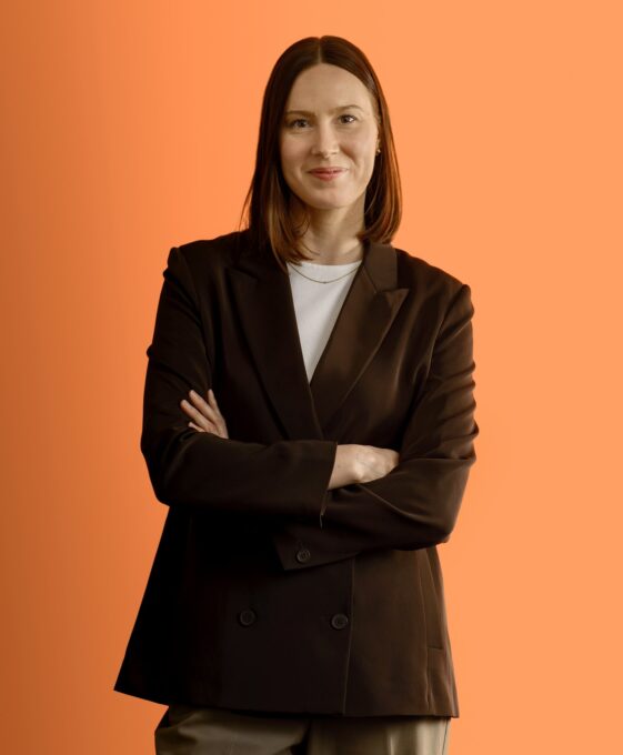 A woman with straight brown hair stands with arms crossed, wearing a black blazer over a white shirt and khaki pants, against an orange background. She is smiling slightly and looking at the camera.