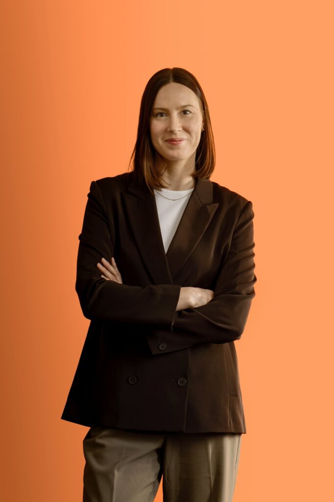 A woman with straight brown hair stands with arms crossed, wearing a black blazer over a white shirt and khaki pants, against an orange background. She is smiling slightly and looking at the camera.