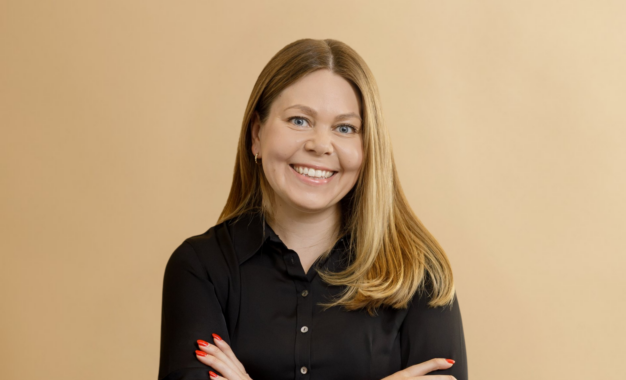 A woman with long blonde hair, wearing a black blouse, smiles with her arms crossed in front of a plain beige background.