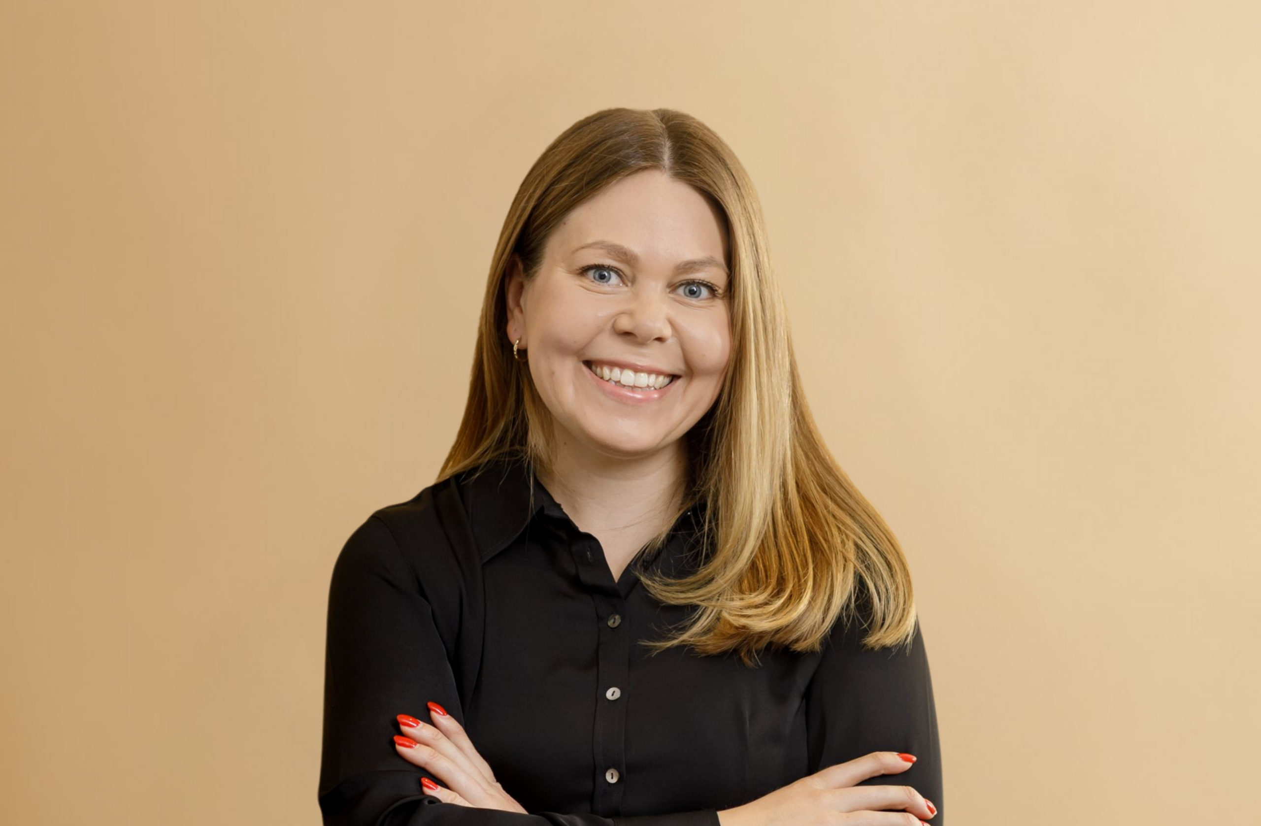 A woman with long blonde hair, wearing a black blouse, smiles with her arms crossed in front of a plain beige background.