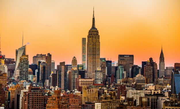The New York City skyline at sunset, with the Empire State Building prominently in the center, surrounded by other skyscrapers against an orange sky.