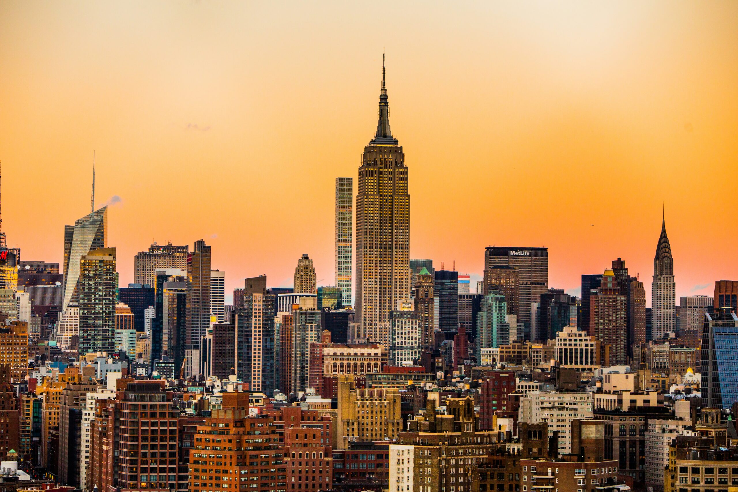 The New York City skyline at sunset, with the Empire State Building prominently in the center, surrounded by other skyscrapers against an orange sky.