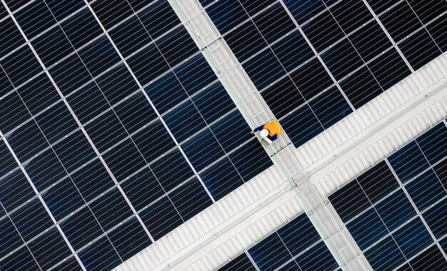 A worker in a yellow vest and helmet walks on a path surrounded by large solar panels, viewed from above.