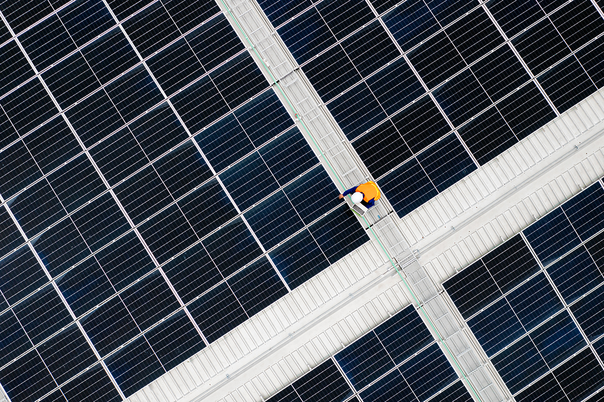 A worker in a yellow vest and helmet walks on a path surrounded by large solar panels, viewed from above.