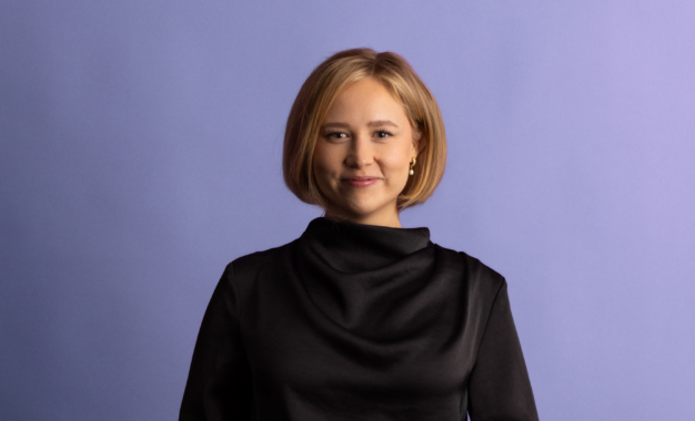 A woman with short blonde hair, wearing a black high-neck top, stands and smiles in front of a plain light purple background.