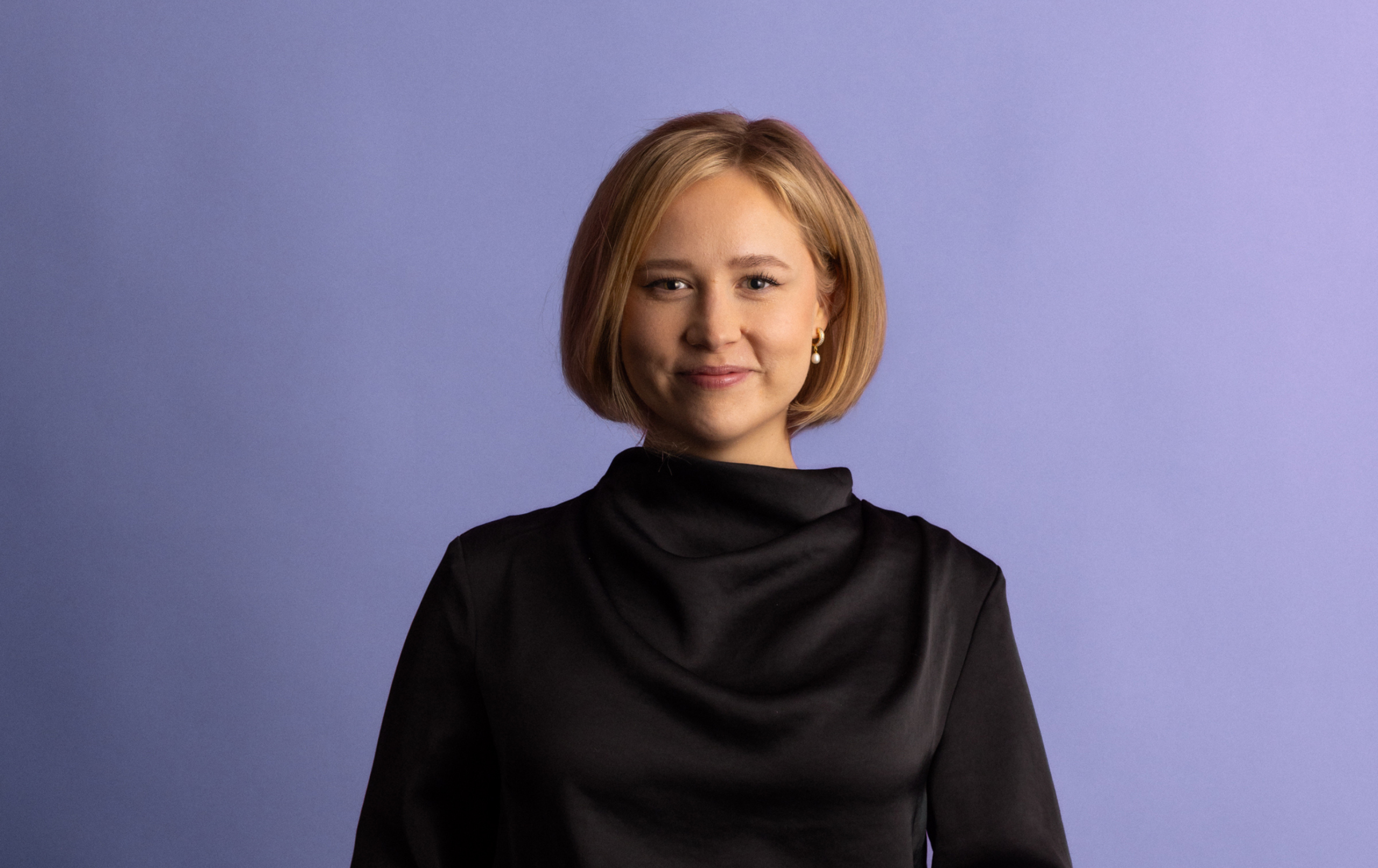 A woman with short blonde hair, wearing a black high-neck top, stands and smiles in front of a plain light purple background.