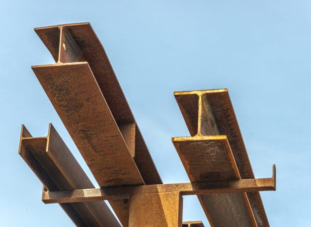 Several rusty steel beams are stacked and angled upwards against a clear blue sky.