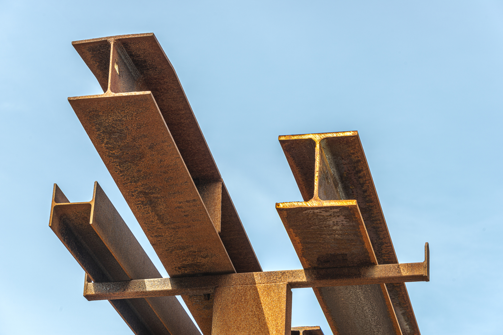 Several rusty steel beams are stacked and angled upwards against a clear blue sky.