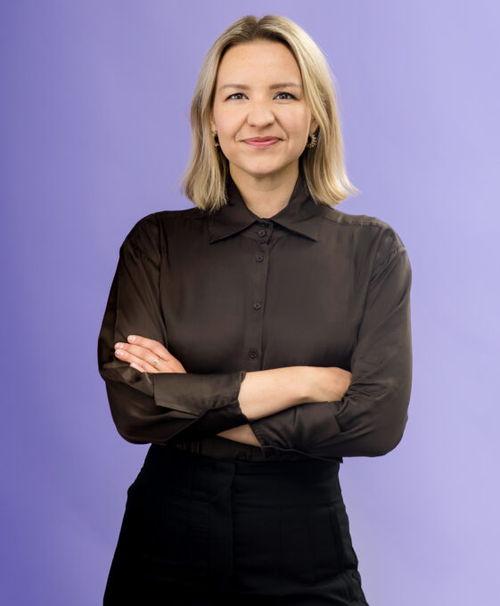 A woman with blonde hair, wearing a black button-up shirt and black pants, stands with arms crossed and smiles in front of a solid light purple background.
