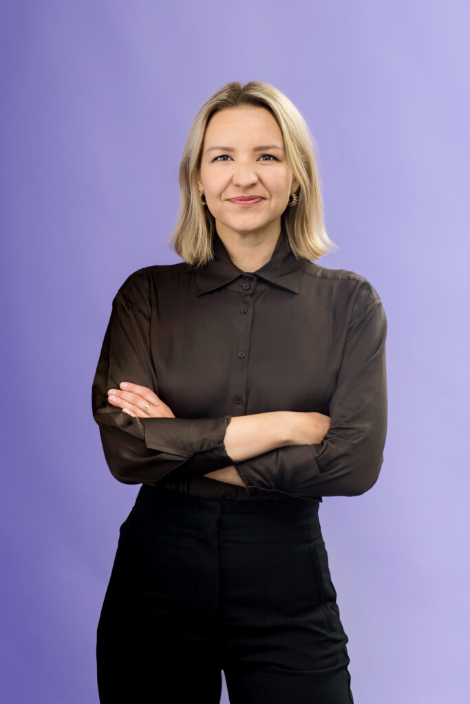 A woman with blonde hair, wearing a black button-up shirt and black pants, stands with arms crossed and smiles in front of a solid light purple background.