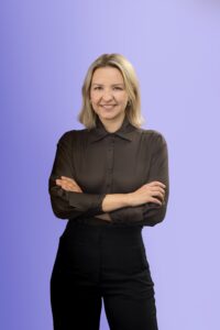 A person with blonde hair, wearing a black sheer blouse and black pants, stands smiling with arms crossed against a light purple background.