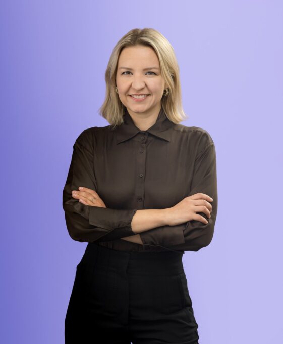 A person with blonde hair, wearing a black sheer blouse and black pants, stands smiling with arms crossed against a light purple background.