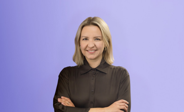 A woman with blonde hair, wearing a sheer black blouse, stands smiling with arms crossed in front of a light purple background.