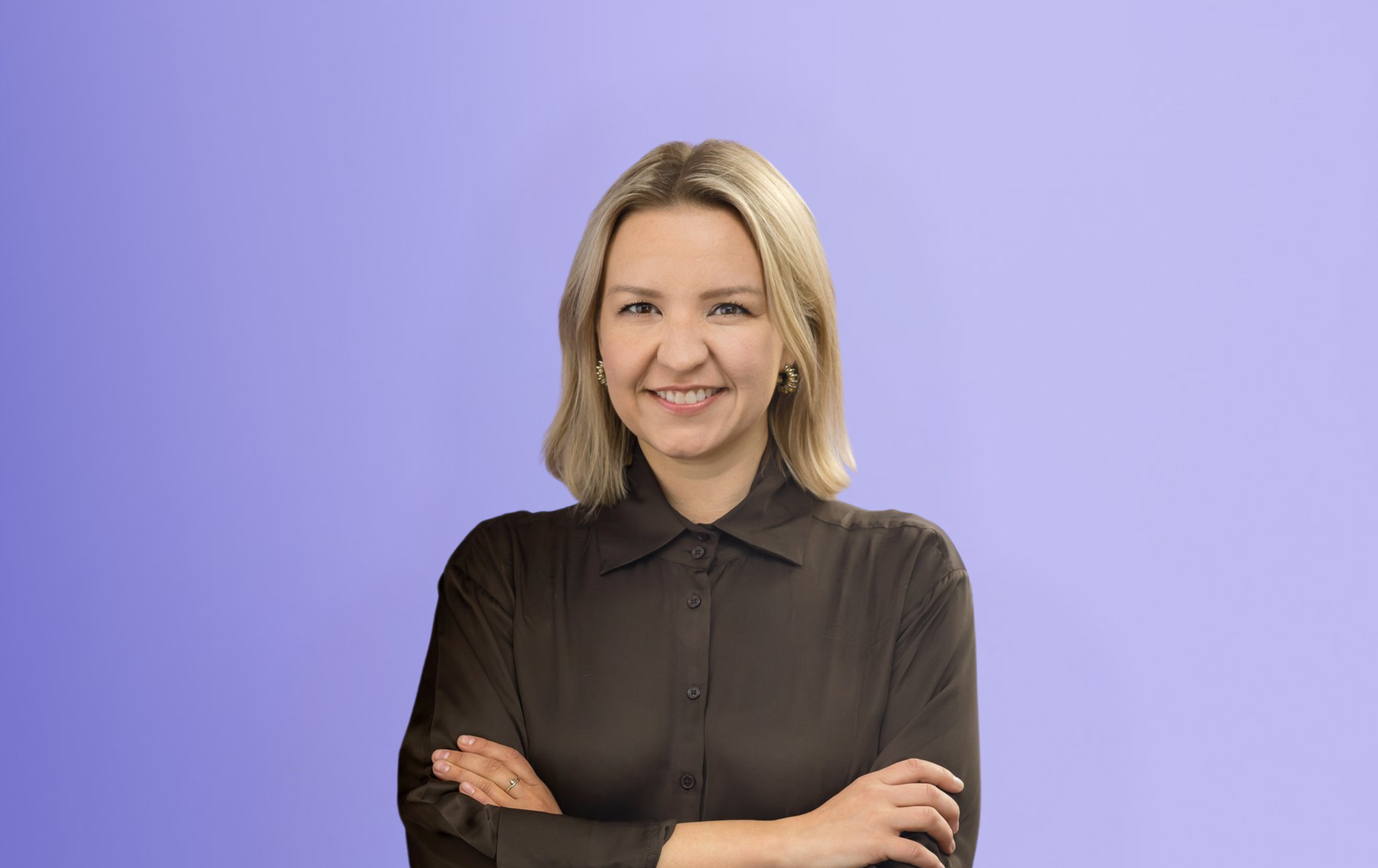 A woman with blonde hair, wearing a sheer black blouse, stands smiling with arms crossed in front of a light purple background.