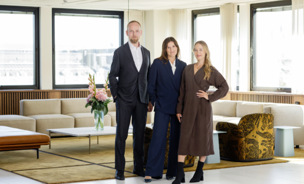 Three people in business attire stand smiling in a modern office lounge with large windows, beige sofas, a yellow rug, and a table with a vase of flowers.
