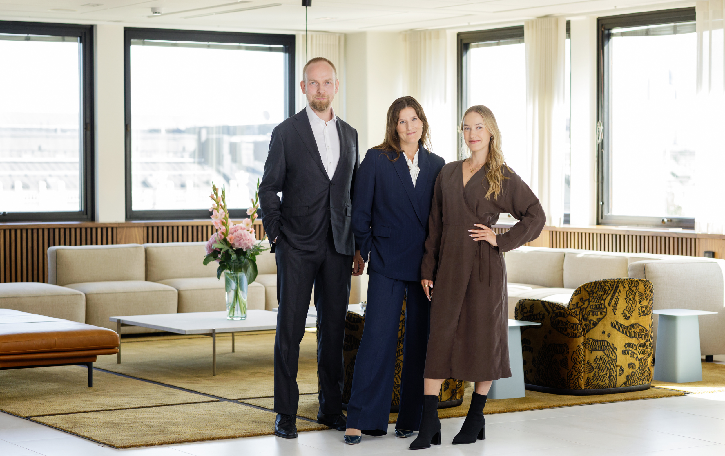 Three people in business attire stand smiling in a modern office lounge with large windows, beige sofas, a yellow rug, and a table with a vase of flowers.