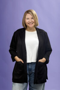 Woman with blonde hair wearing a black blazer, white shirt, and jeans stands smiling with hands in her pockets against a light purple background.