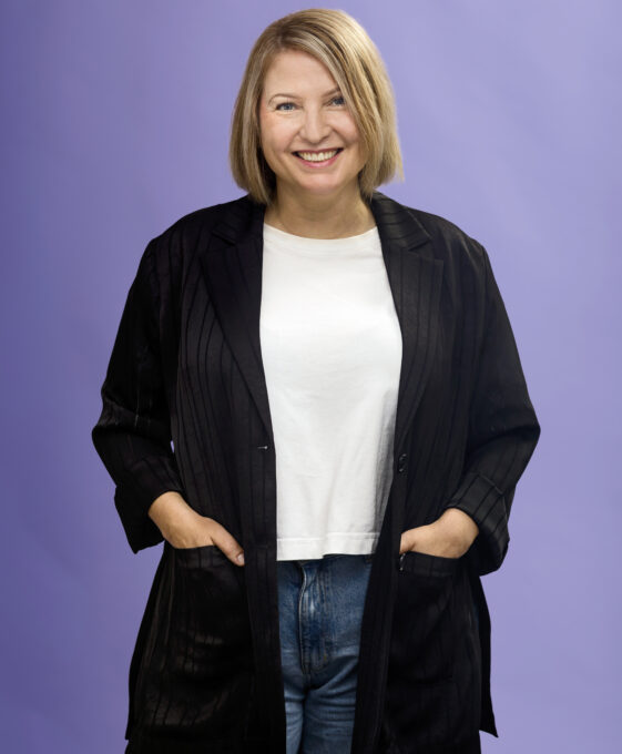 Woman with blonde hair wearing a black blazer, white shirt, and jeans stands smiling with hands in her pockets against a light purple background.