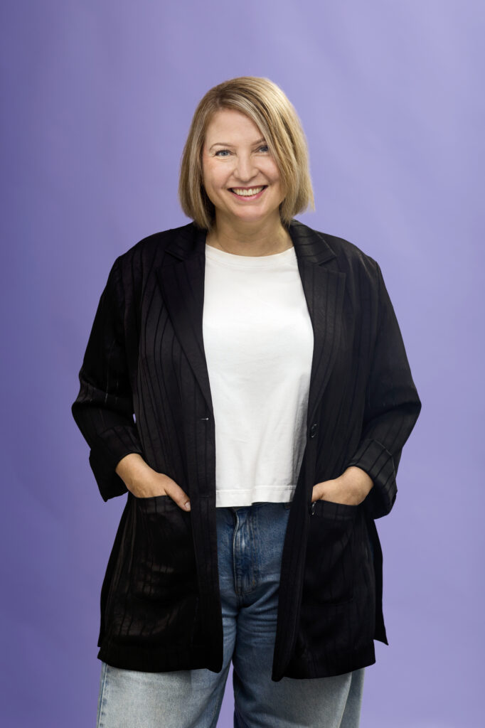 Woman with blonde hair wearing a black blazer, white shirt, and jeans stands smiling with hands in her pockets against a light purple background.